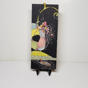 May include: A vintage bookmark with a black background and a colorful illustration of a woman in a pink dress and a red hat, surrounded by clouds and a heart shape. The bookmark has a yellow string attached to the top.