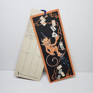 May include: Vintage bookmark with a devil illustration. The bookmark features an orange devil figure with a pitchfork, surrounded by playing cards on a black background with an orange border. A second, cream-colored card is also present.