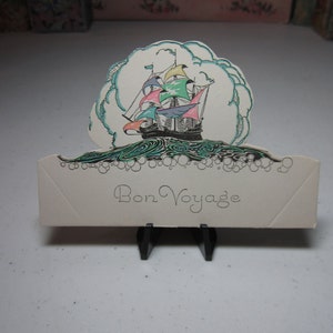 May include: A white card with the text "Bon Voyage" printed in black cursive script. The card is decorated with a colorful illustration of a ship sailing on a blue sea with white clouds.
