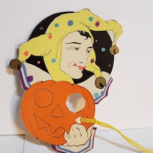May include: A vintage Halloween decoration featuring a jester with a yellow hat and a pumpkin with a carved face. The jester is holding the pumpkin with a string attached to the pumpkin's mouth.