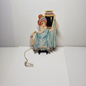 May include: A vintage paper doll cutout of a woman in a blue robe holding a black and gold vase. The doll is attached to a string.