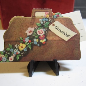 May include: A brown paper suitcase with a floral garland and a tag that says "Greetings". The suitcase is on a black stand.