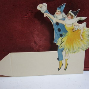 May include: A vintage paper cutout featuring a couple in theatrical costumes. The man wears a blue outfit with black accents, while the woman is in a yellow dress. The cutout is attached to a cream-colored base.