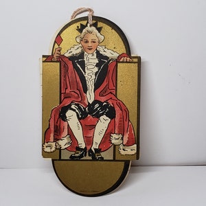 May include: A vintage card-shaped paper ornament featuring a king in a red and black robe sitting on a throne. The king is holding a red playing card with a diamond symbol. The ornament is gold with a white border.