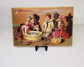 Könnte beinhalten: Eine rechteckige Vintage-Postkarte mit dem Schriftzug "Hallowe'en" in roter Schreibschrift. Die Illustration zeigt drei Kinder in historischer Kleidung: eines beim Apfeltauchen in einem Holzzuber, eines sitzend und eines neben einem rauchenden Kürbis.