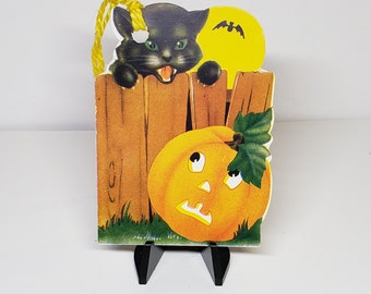 May include: Vintage Halloween decoration featuring a black cat with green eyes peeking over a wooden fence. A yellow moon with a bat silhouette and an orange pumpkin with a carved face complete the festive scene. The decoration is mounted on a black stand.