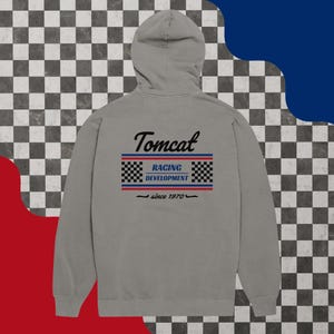May include: A gray hoodie with a hood, featuring the text "Tomcat" above a racing-themed design with the words "Racing Development" and "since 1970". The design includes checkered flag patterns and red and blue stripes.