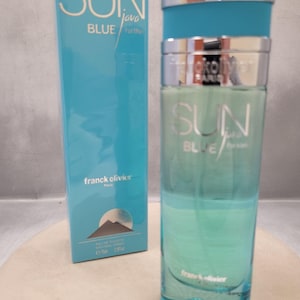 Puede incluir: Un frasco de vidrio cilíndrico y alto de agua de tocador Sun Java Blue con líquido azul claro, una tapa plateada y una caja rectangular azul claro a juego que presenta el texto Sun Java Blue, Franck Olivier Paris y un gráfico de una montaña.