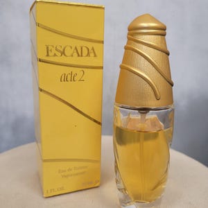 Puede incluir: Un frasco de Eau de Toilette Escada Acte 2 con tapón dorado y caja amarilla a juego. La caja presenta el logotipo de Escada y el nombre del producto. El frasco contiene un líquido de color ámbar claro y está etiquetado como 30 ml.
