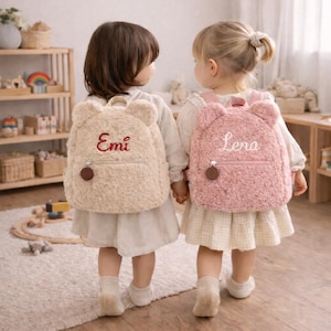 Può includere: Due bambini con zaini a forma di orsetto. Uno è beige con "Emi" ricamato in rosso, l'altro è rosa con "Lena" ricamato in bianco. Entrambi hanno una cerniera e un dettaglio circolare marrone.