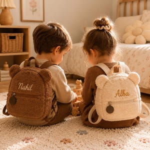 Könnte beinhalten: Zwei kleine Teddybär-Rucksäcke aus weichem, flauschigem Sherpa-Material. Ein Rucksack ist dunkelbraun mit dem in Weiß gestickten Namen Nahil, der andere ist cremefarben mit dem in Braun gestickten Namen Alba. Beide verfügen über Bärenohren.