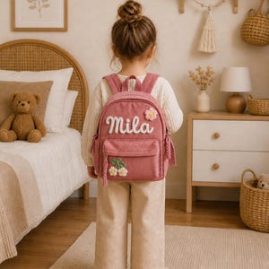 Peut inclure: Un sac à dos en velours côtelé rose poudré portant le prénom Mila en lettres cursives blanches, orné de petites applications de fleurs blanches et jaunes sur la poche avant et le coin supérieur, avec des bretelles et des pompons assortis.