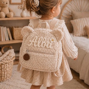 Op de afbeelding: Een beige rugzak in de vorm van een teddybeer met de naam "Alba" op de voorkant geborduurd. De rugzak heeft een klein ritsvak en een bruin rond label. Het kind draagt een beige jurk met een geruite rok.