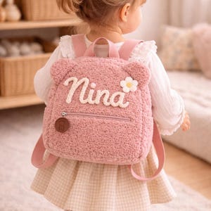 Puede incluir: Una mochila rosa de felpa con el nombre "Nina" bordado en la parte delantera. La mochila tiene orejas de oso, una pequeña flor y un bolsillo con cremallera. Las correas y un pequeño dije también son rosas.