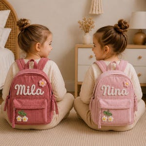 Peut inclure: Deux sacs à dos en velours côtelé, l'un rose foncé et l'autre rose clair, chacun doté d'une poche avant, d'une petite fleur brodée et d'une étiquette nominative Mila ou Nina en lettres cursives blanches, avec des tirettes tressées.