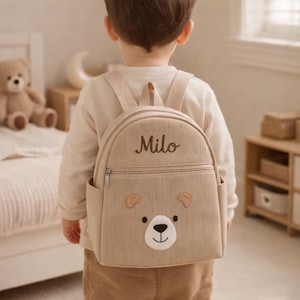 Peut inclure: Un sac à dos en velours côtelé beige avec un motif de visage d'ours sur la poche avant et le prénom Milo brodé en écriture cursive marron foncé. Le sac possède deux bretelles, une poignée supérieure et un compartiment principal zippé.
