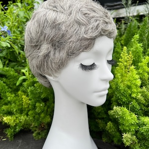 May include: A mannequin head with a short, gray wig. The wig has a textured, layered style. The mannequin has long, black eyelashes and closed eyes. The background is filled with green foliage.