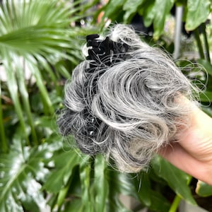 Könnte beinhalten: Eine Haarklammer mit einem synthetischen Haarteil in einer Mischung aus Grau-, Silber- und Schwarztönen. Das Haar ist zu einem voluminösen, welligen und strukturierten Dutt geformt und sicher an einer schwarzen Kunststoffklammer mit Zinken befestigt.