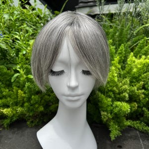 May include: A mannequin head with a short, layered wig. The wig is a mix of gray and light brown, styled with a side part and bangs. The mannequin has closed eyes and long, dark eyelashes. The background is a lush green garden.