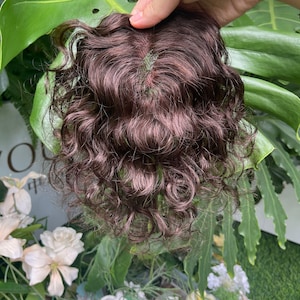 May include: A dark brown hair topper featuring a wavy, curly texture and a visible monofilament base at the crown. The hairpiece has a natural-looking part with a skin-coloured base, designed to add volume and coverage to the top of the head.