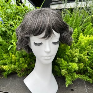 May include: A short, gray wig with dark roots and curled ends is displayed on a white mannequin head. The wig has a layered style with bangs. The mannequin has long, dark eyelashes and is set against a backdrop of green foliage.
