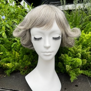 May include: A mannequin head with a short, gray wig. The wig has a side part and is styled with soft waves. The mannequin has long, black eyelashes and is set against a backdrop of green foliage.