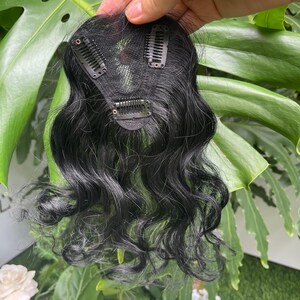 May include: A black hair topper featuring a mesh base with three attached metal pressure-sensitive clips. The hair is wavy and dark, extending from the base in a natural-looking style. The underside of the hairpiece shows the construction.