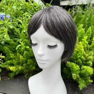 May include: A short, gray wig with dark roots is styled on a white mannequin head. The wig has a natural-looking hairline and is surrounded by green foliage. The mannequin has long, dark eyelashes.