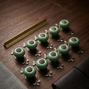 May include: A collection of ten green ceramic cabinet knobs with bronze-colored floral accents. The knobs are arranged on a dark wooden surface, with a brass ruler and a dark leather notebook visible in the background. The knobs are a decorative hardware item.