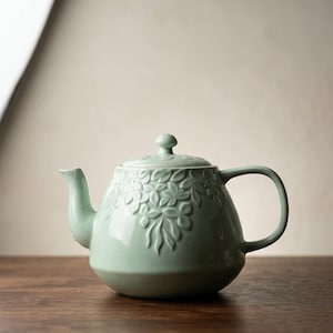 May include: A light green ceramic teapot with a floral design. The teapot has a curved spout, a rounded body, and a handle. The lid has a small knob. The teapot is sitting on a wooden surface.