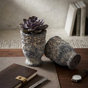 May include: Two decorative, weathered-look ceramic planters with ornate scrollwork. One upright planter holds a purple succulent. A brown leather-bound notebook, a silver pen, and a wristwatch with a brown leather strap are also on the table.