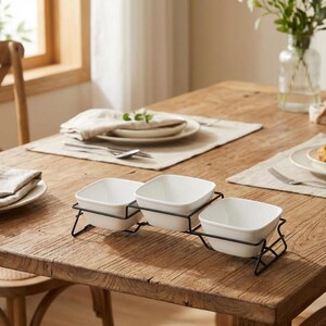 May include: A serving dish set with three white square bowls in a black metal rack. The bowls are arranged in a row on a wooden table, with place settings and a vase of flowers in the background. The set is designed for serving appetizers or snacks.