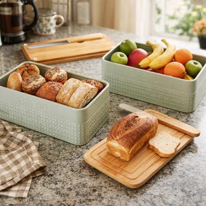 May include: A kitchen scene with bread and fruit. Two rectangular, light green containers hold bread and fruit. A wooden cutting board with a loaf of bread and a knife is in the foreground. A wooden cutting board with a knife is in the background.