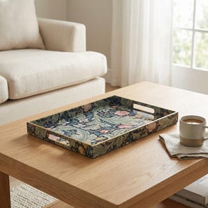 May include: Rectangular decorative tray with a floral pattern in shades of blue, green, and pink. The tray has gold-colored edges and cut-out handles. It sits on a light wood coffee table, next to a mug and a folded linen napkin.
