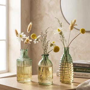 May include: Three decorative glass vases with dried flowers. The vases are green and gold, with textured and embossed designs. The vases are filled with water and contain various dried flowers, including daisies and yellow globe amaranth.
