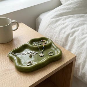 May include: A green ceramic jewelry tray with an organic shape. The tray holds various pieces of jewelry, including necklaces, rings, and a bracelet. The tray is placed on a light-colored wooden surface, with a mug visible in the background.