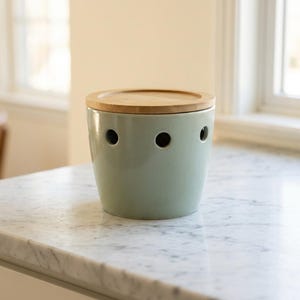 May include: A light green ceramic garlic keeper with a wooden lid. The keeper has three circular ventilation holes and sits on a marble countertop. The container is cylindrical and has a smooth, glossy finish.