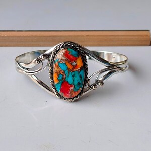 Könnte beinhalten: Ein silberfarbenes Armreif-Armband mit einem ovalen, mehrfarbigen Stein. Der Stein weist ein Mosaik aus Türkis-, Rot-, Orange- und Brauntönen auf. Das Armband hat einen dekorativen, seilartigen Rand um den Stein.
