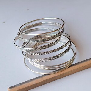 Puede incluir: Un conjunto de pulseras de plata, algunas con intrincados diseños florales y de hojas, y otras con un diseño liso y sencillo. Las pulseras se exhiben sobre una superficie blanca con una herramienta de madera en primer plano.