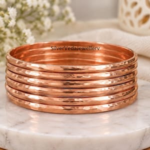 May include: A stack of five circular copper bangles with a hammered, textured finish. The bangles have a warm, metallic reddish-brown color and a polished surface. The text SilverVedaJewelleryv is visible on the top edge of the stack.