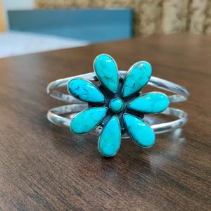 Könnte beinhalten: Ein silbernes Armband mit einem zentralen Blumenmotiv, das aus sechs tropfenförmigen Türkissteinen und einem kleinen runden Türkisstein in der Mitte besteht. Das Metallband ist glatt und poliert und bildet eine offene, kreisförmige Form.