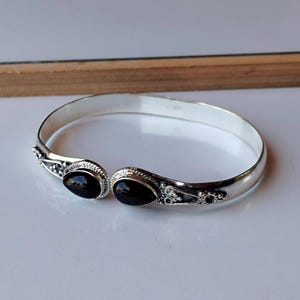 Può includere: Un bracciale a polsino color argento con due pietre preziose nere ovali. Il bracciale presenta dettagli decorativi attorno alle pietre preziose e una fascia semplice e lucida. Le pietre preziose misurano circa 1,3 cm di lunghezza.