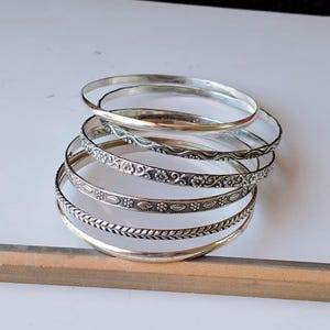 May include: A stack of silver bangles with various designs. Some bangles are plain, while others feature floral, leaf, and patterned details. The bangles are displayed on a white surface.
