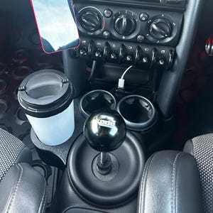 Könnte beinhalten: Auto-Innenraum mit einem schwarzen Schalthebelknauf mit dem Wort "Kode" in Weiß, einem weiß-schwarzen Reisebecher und einem Telefon. Das Armaturenbrett hat verschiedene Bedienelemente und Getränkehalter. Die Autositze sind schwarz.