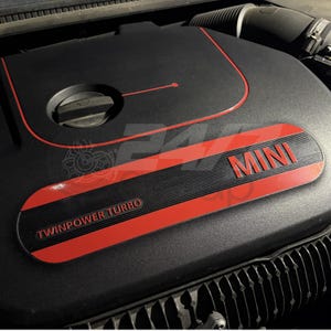 May include: A black engine cover for a vehicle featuring a red accent line and a red horizontal plate with the text MINI and TWINPOWER TURBO. The cover includes a circular oil filler cap and a textured surface with ribbed detailing on the side.