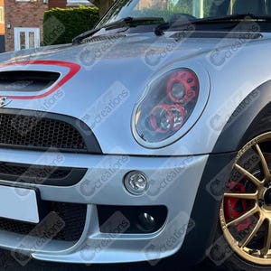 Peut inclure: Mini Cooper argenté avec des détails rouges sur le capot et les phares. La voiture a une calandre noire, un pare-chocs avant et des roues dorées. Les phares ont des détails rouges.