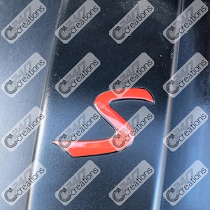 Peut inclure: Gros plan d'un emblème "S" rouge brillant sur une surface gris foncé. L'emblème a un design incurvé. L'image contient plusieurs instances du texte "347 creations".