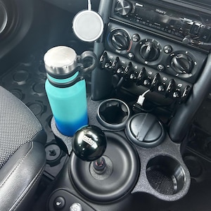 May include: Car interior showing a blue water bottle in a cup holder. A black gear stick with white text is visible, along with a wireless charger and a car stereo.