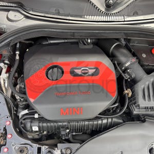 May include: A detailed shot of a Mini Cooper engine bay. The engine cover is predominantly black with a red section. The word "MINI" is in red, alongside the "MINI" logo. The words "TWINPOWER TURBO" are also visible on the engine.