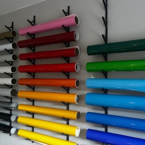 May include: A display of various colored vinyl rolls, including pink, red, orange, yellow, green, and blue. The rolls are mounted on black metal racks, showcasing a range of colors for crafting or design projects. The rolls are of varying lengths.